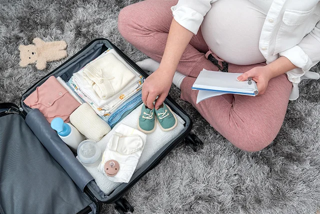 Pregnant woman sitting on a bed packing baby clothes into a hospital bag before delivery.